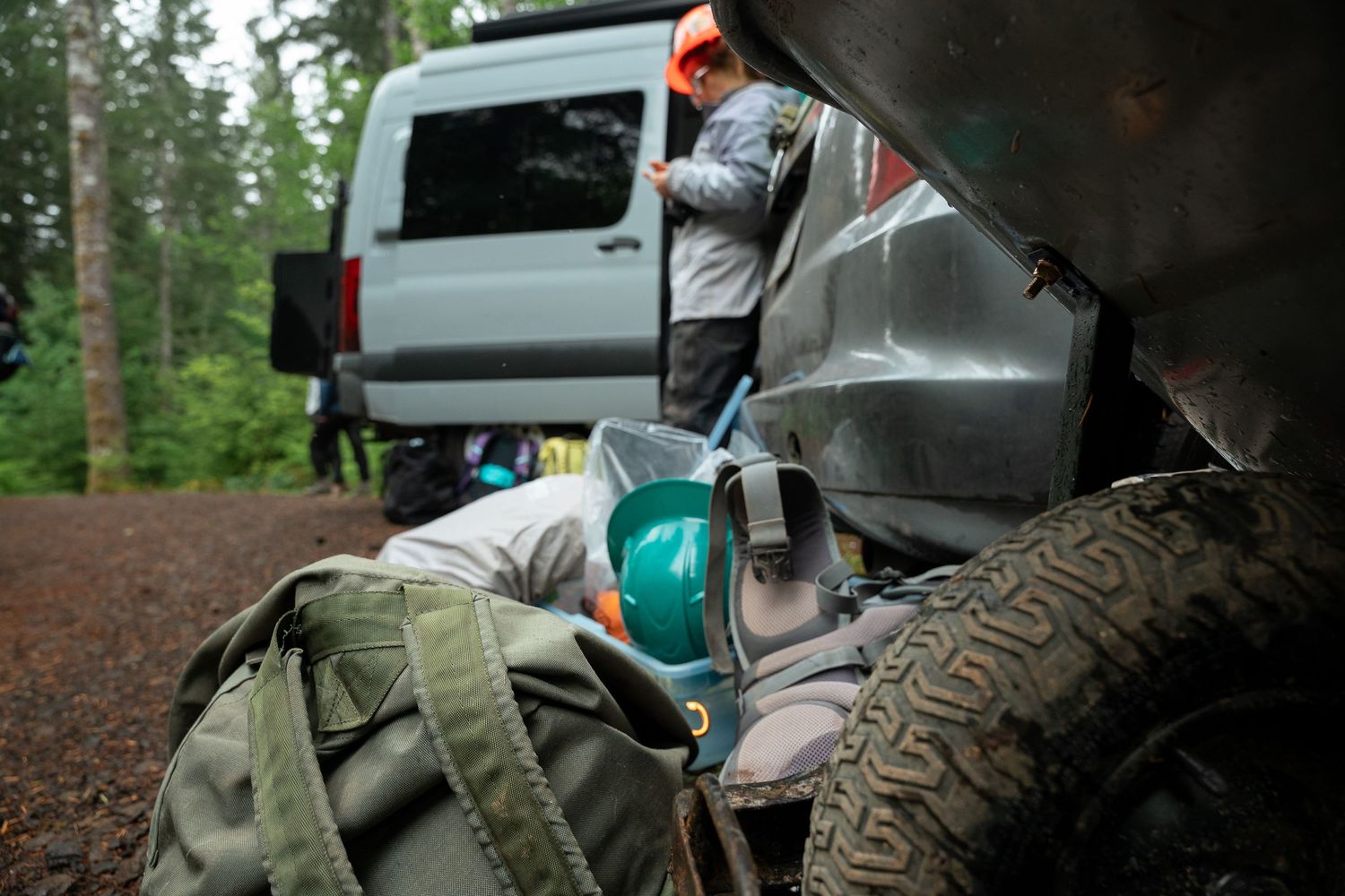 Outdoor gear and backpack near an ATV in a forest campsite with a person standing by a van.