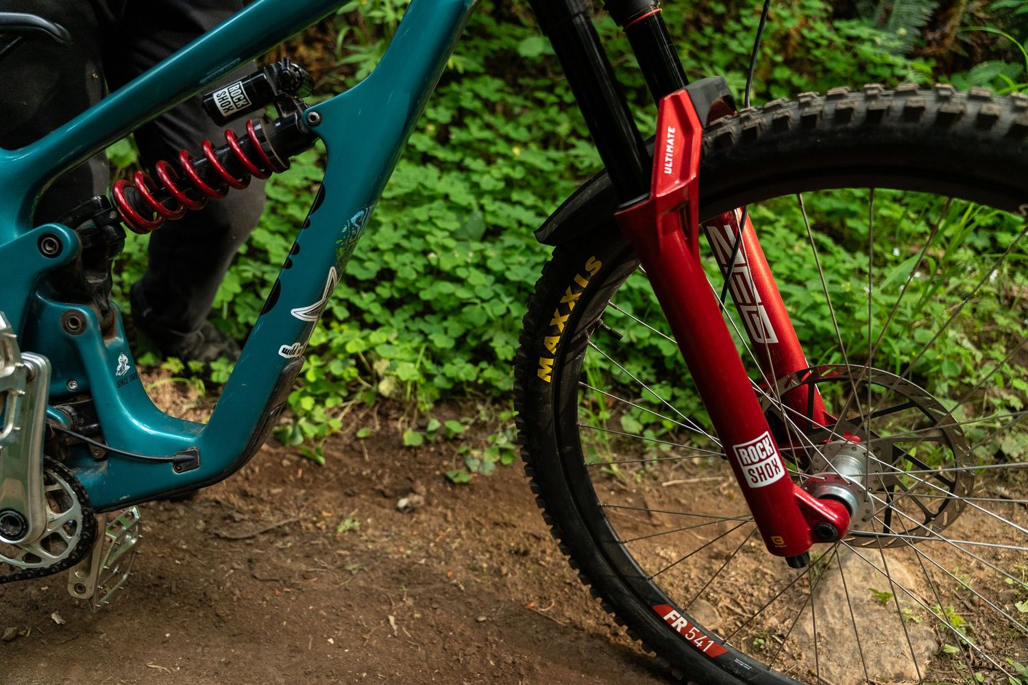 Close-up of a teal mountain bike with red suspension fork and rear shock on a forest trail.