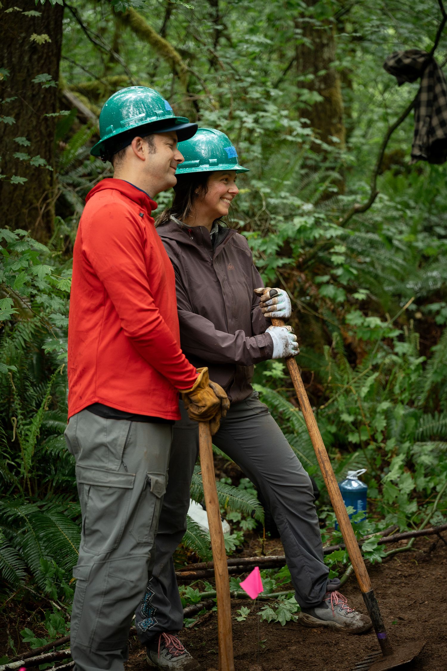 Two volunteers in green hard hats and work gloves use shovels for trail maintenance in a lush forest setting.