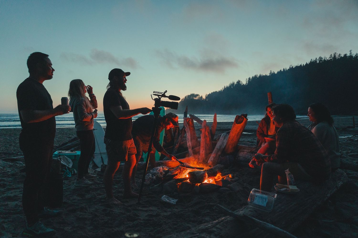 Group of friends gathered around a campfire on a beach at dusk, enjoying an outdoor bonfire by the ocean.