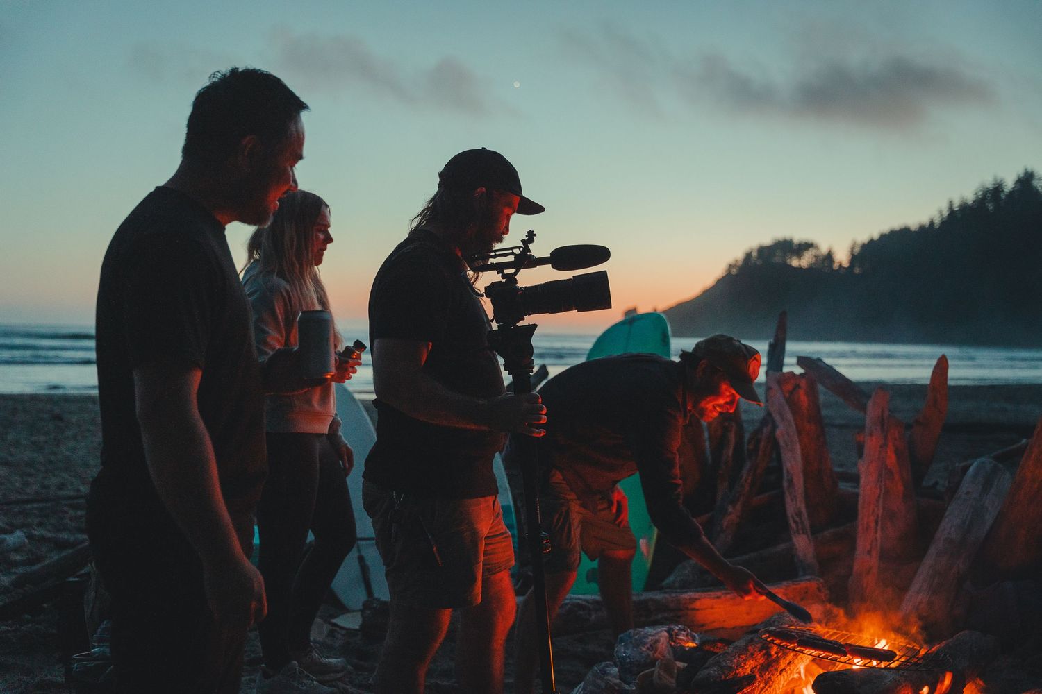 Group of people filming with a camera by a beach campfire at dusk with ocean and trees in background.