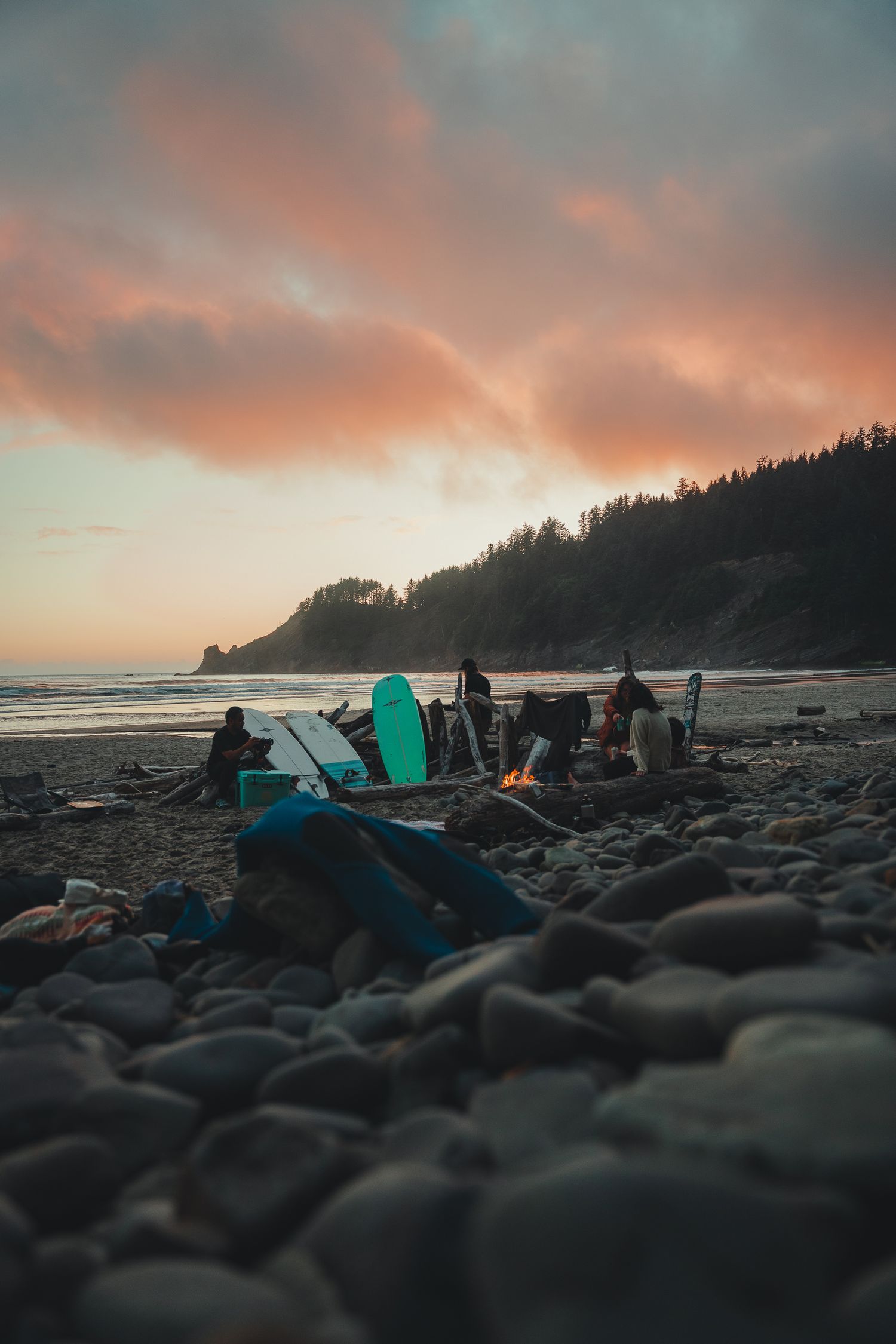 Surfers gather around a beach bonfire at sunset with surfboards on rocky shore, colorful pink clouds over ocean.