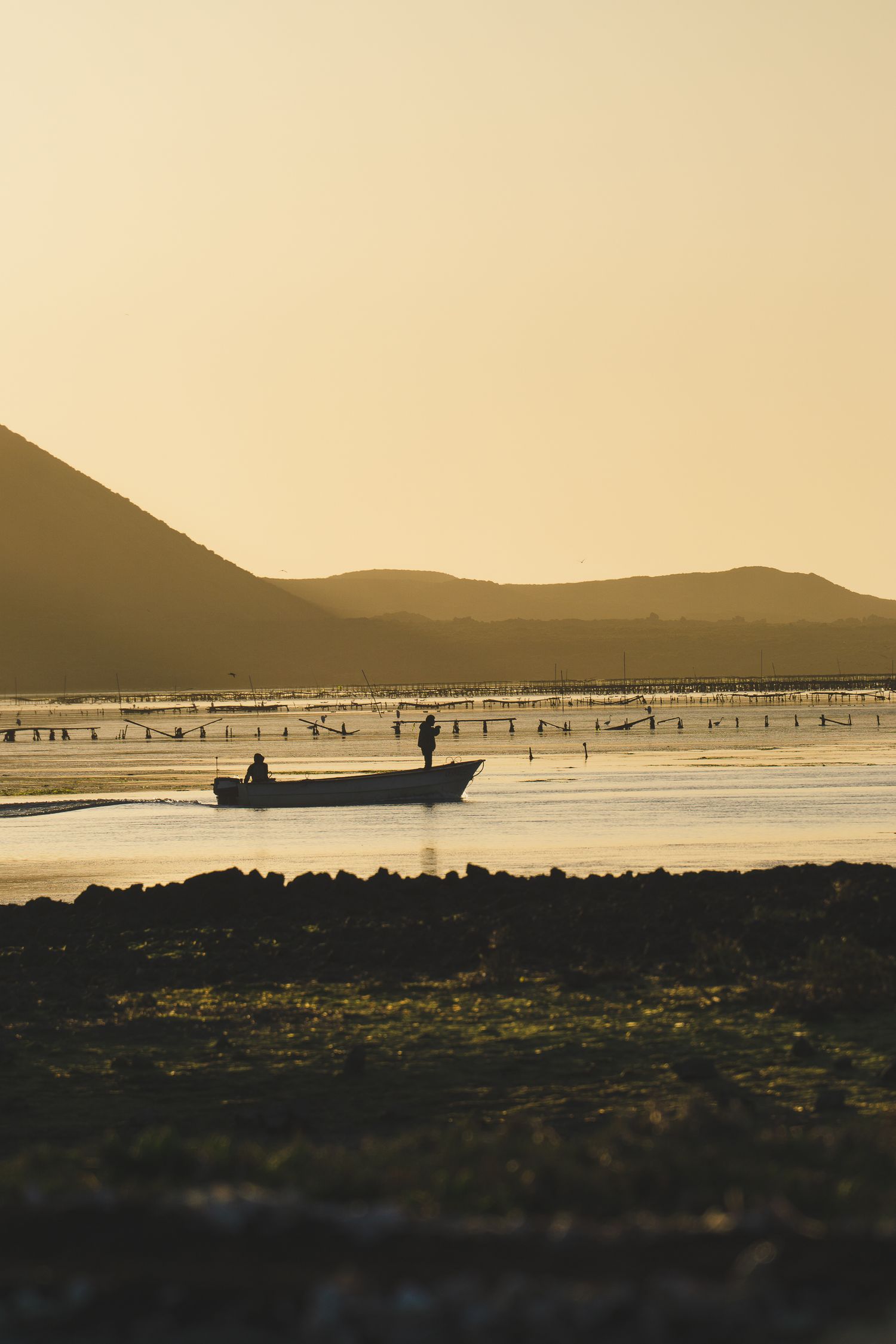 Silhouette of a small fishing boat with two people gliding across calm golden waters at sunset with mountains in background.