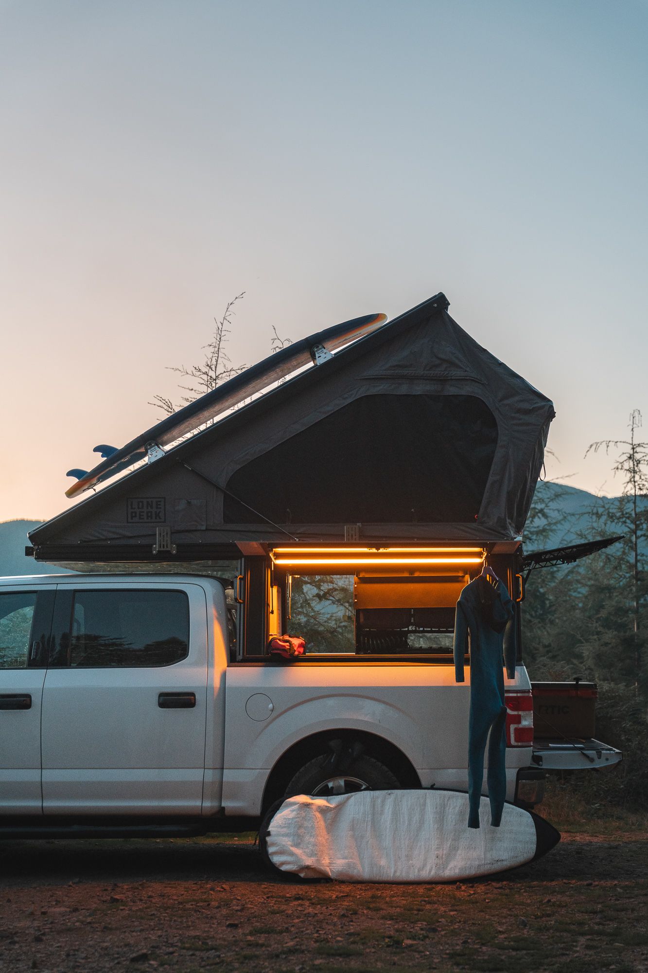 Side view of illuminated rooftop tent setup on white pickup truck at dusk with warm lighting.