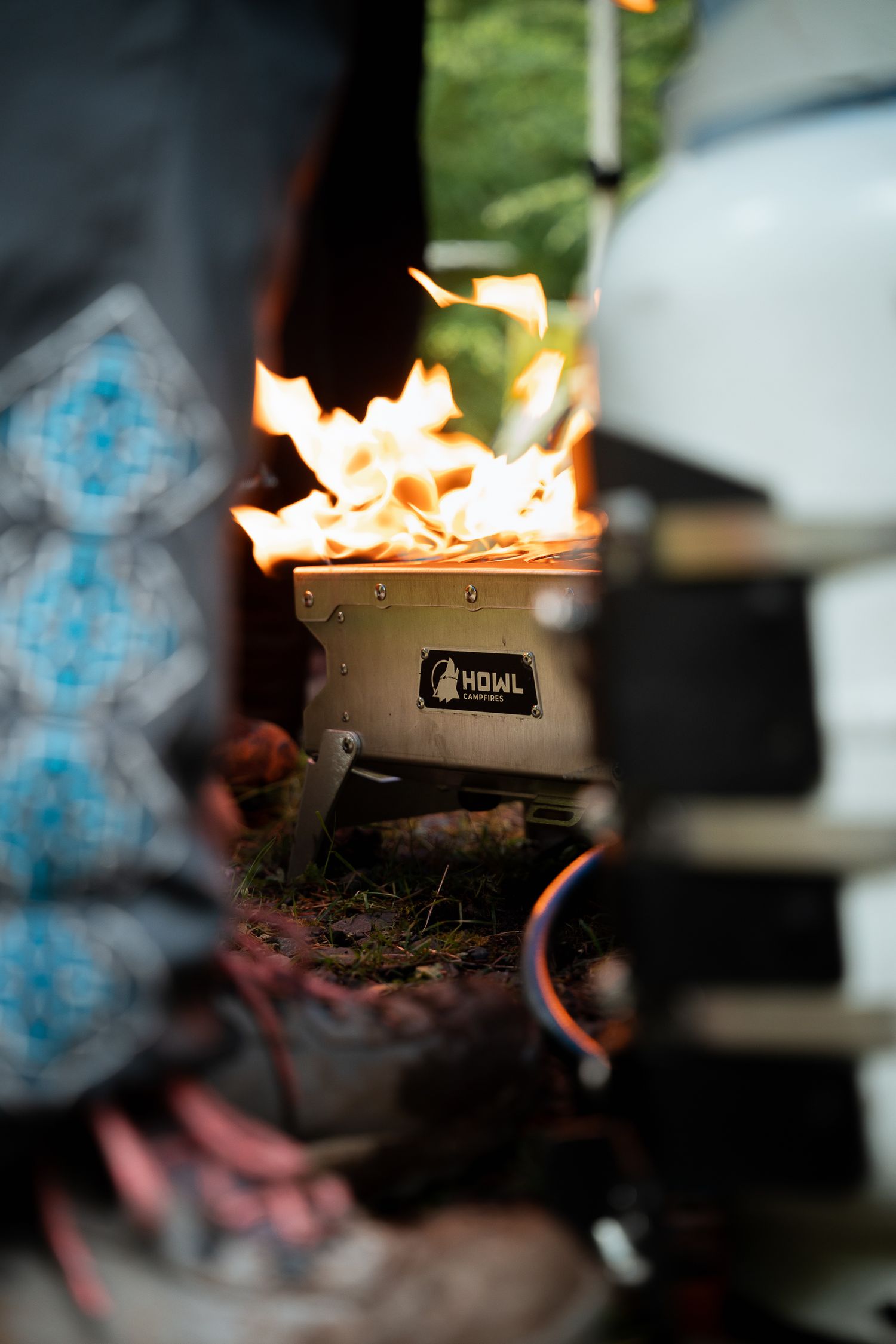 HOWL portable camp stove burning with bright flames outdoors near a vehicle, surrounded by forest gear.