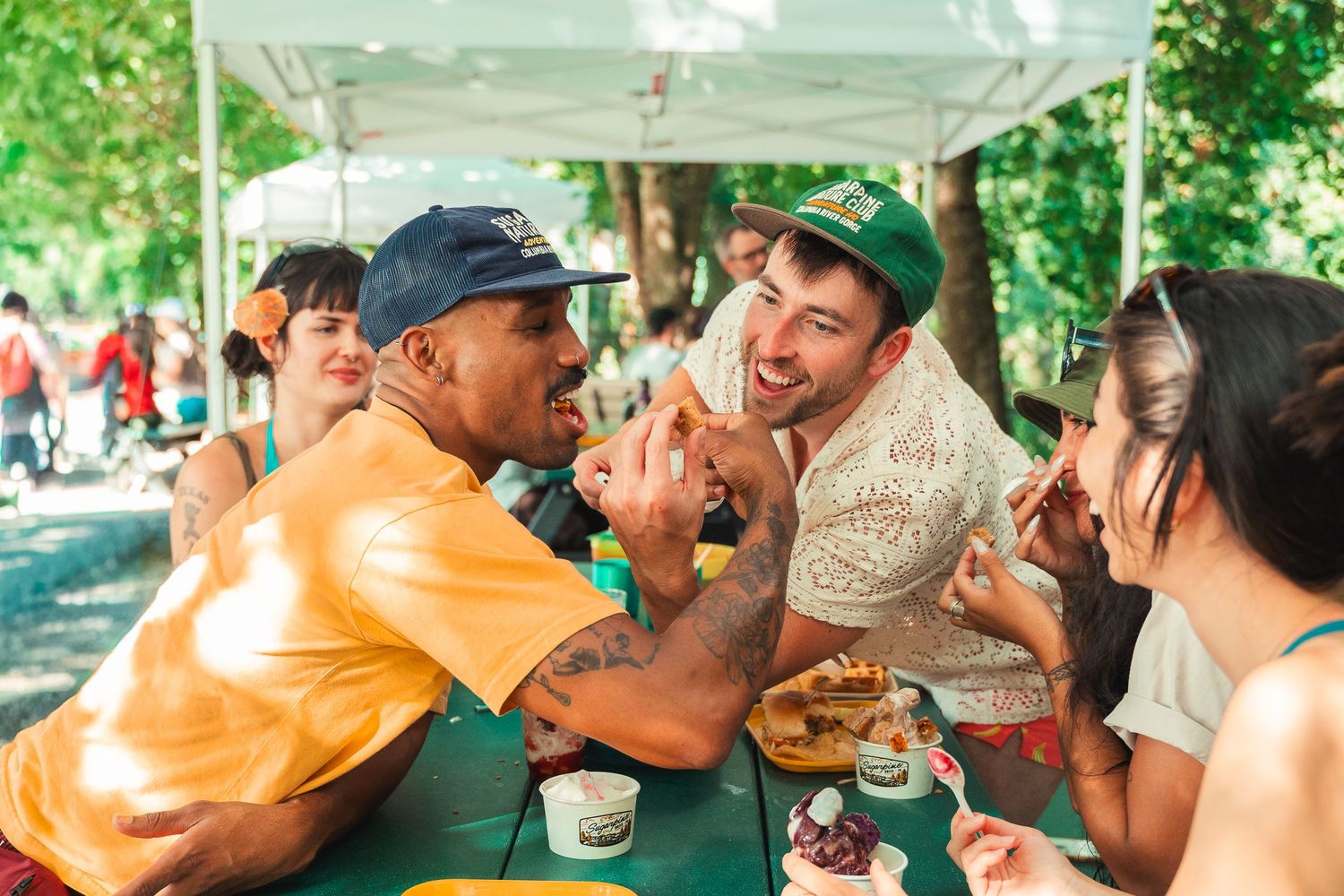 Friends laughing and sharing food at an outdoor picnic table under green trees at a summer festival.