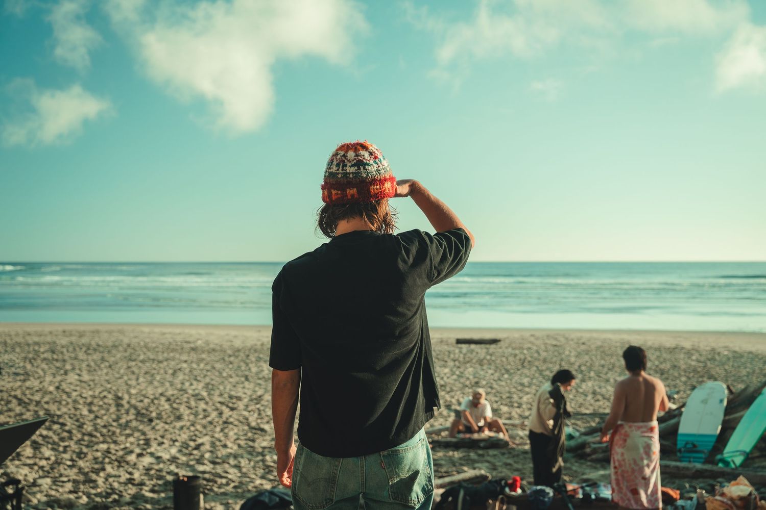 Person wearing a bandana photographing the ocean at a sunny beach with surfers and beachgoers in the background.