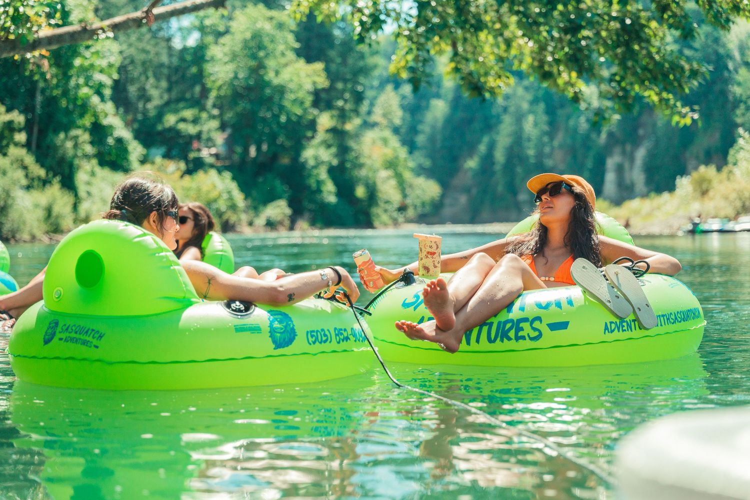Two women relaxing on green inflatable tubes floating on a calm river on a sunny day.