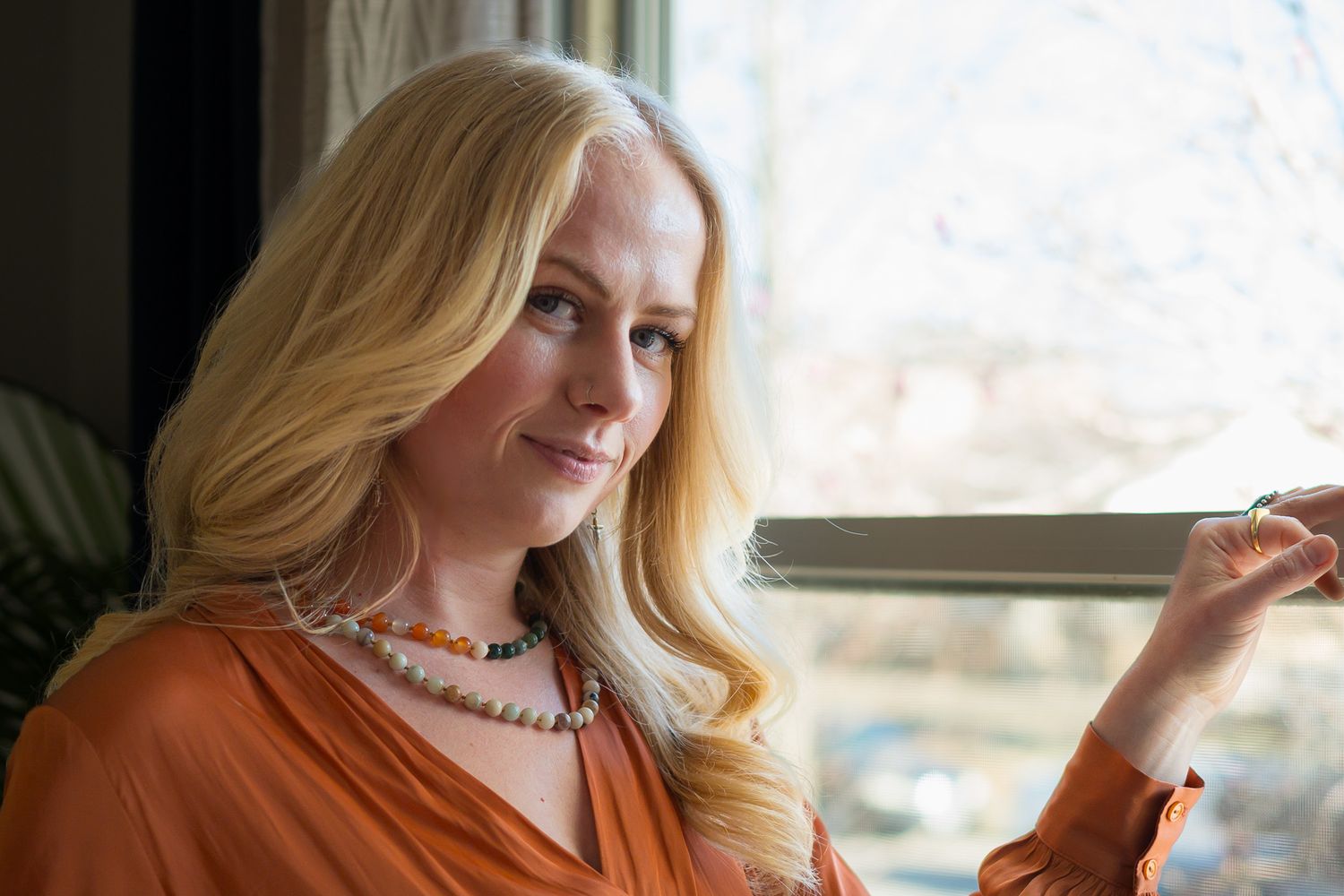 Smiling blonde woman in orange top wearing a beaded necklace near a sunlit window.