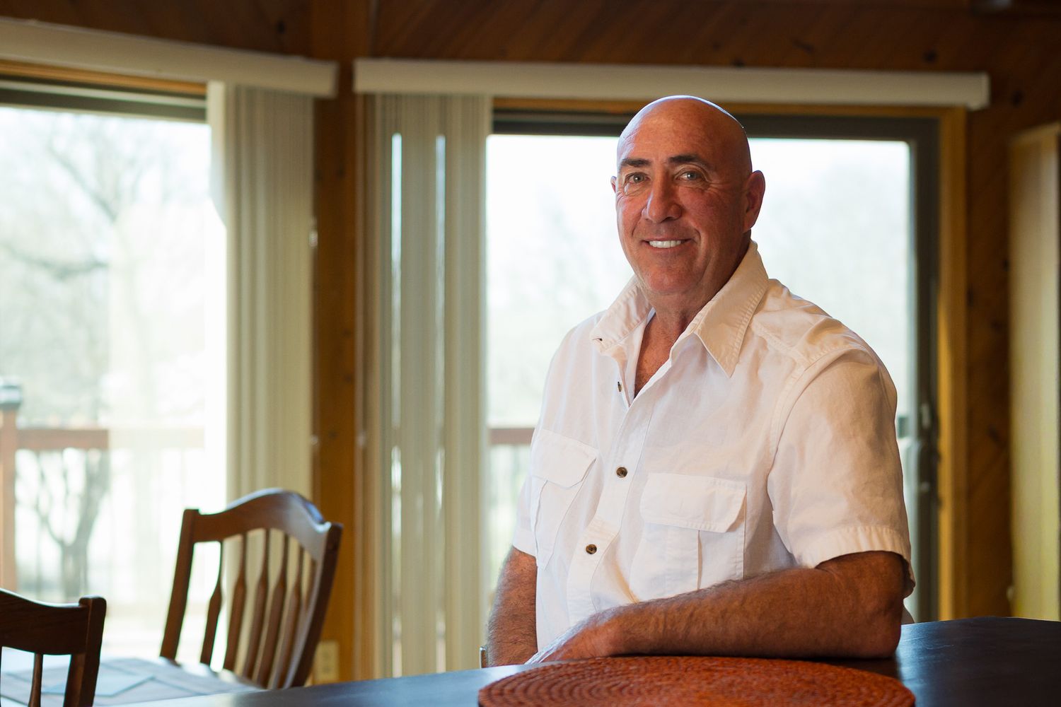 Smiling middle-aged man in white shirt sitting at dining table near large windows in a cozy home.