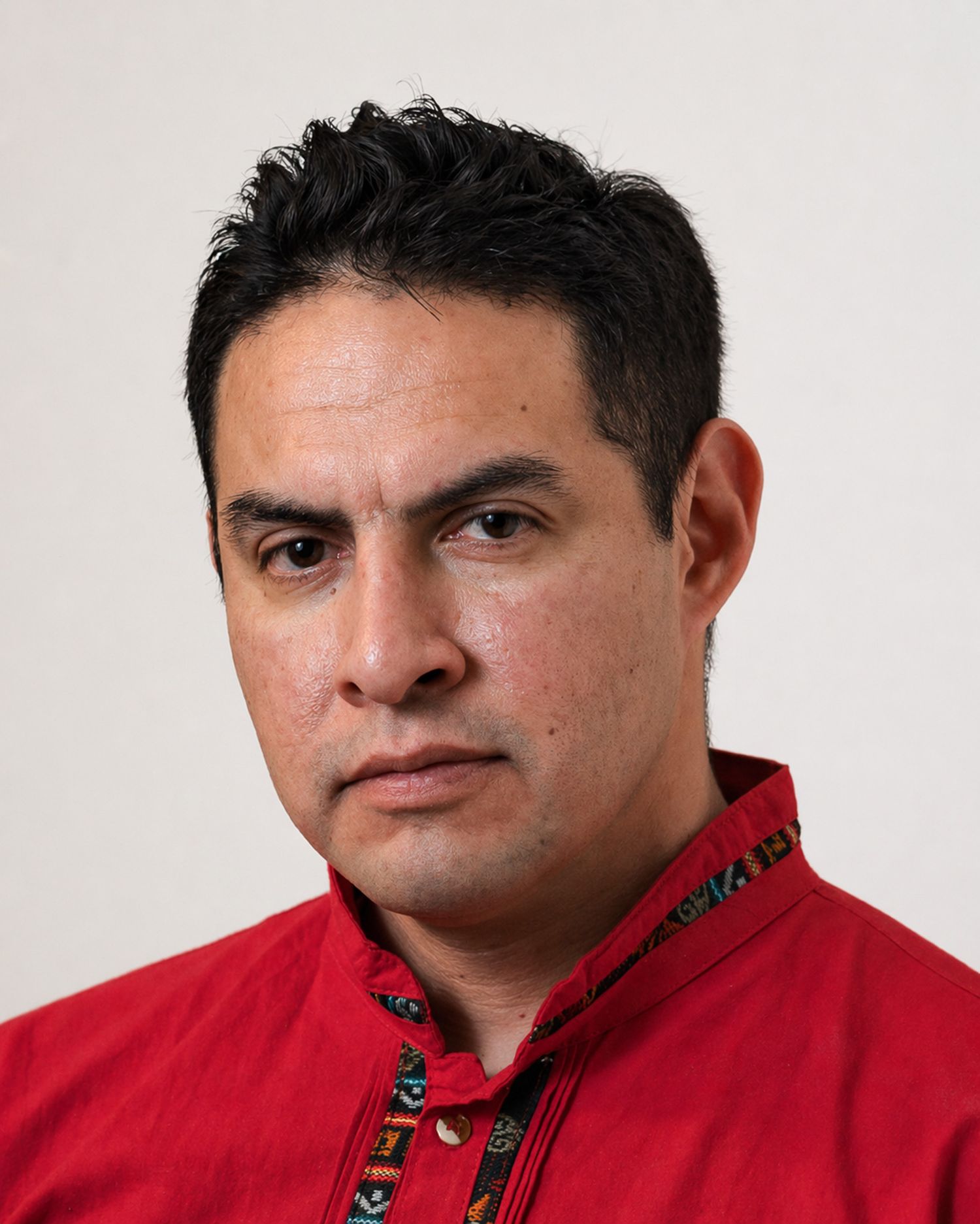 Man in red embroidered shirt with serious expression poses for a formal portrait against neutral background.