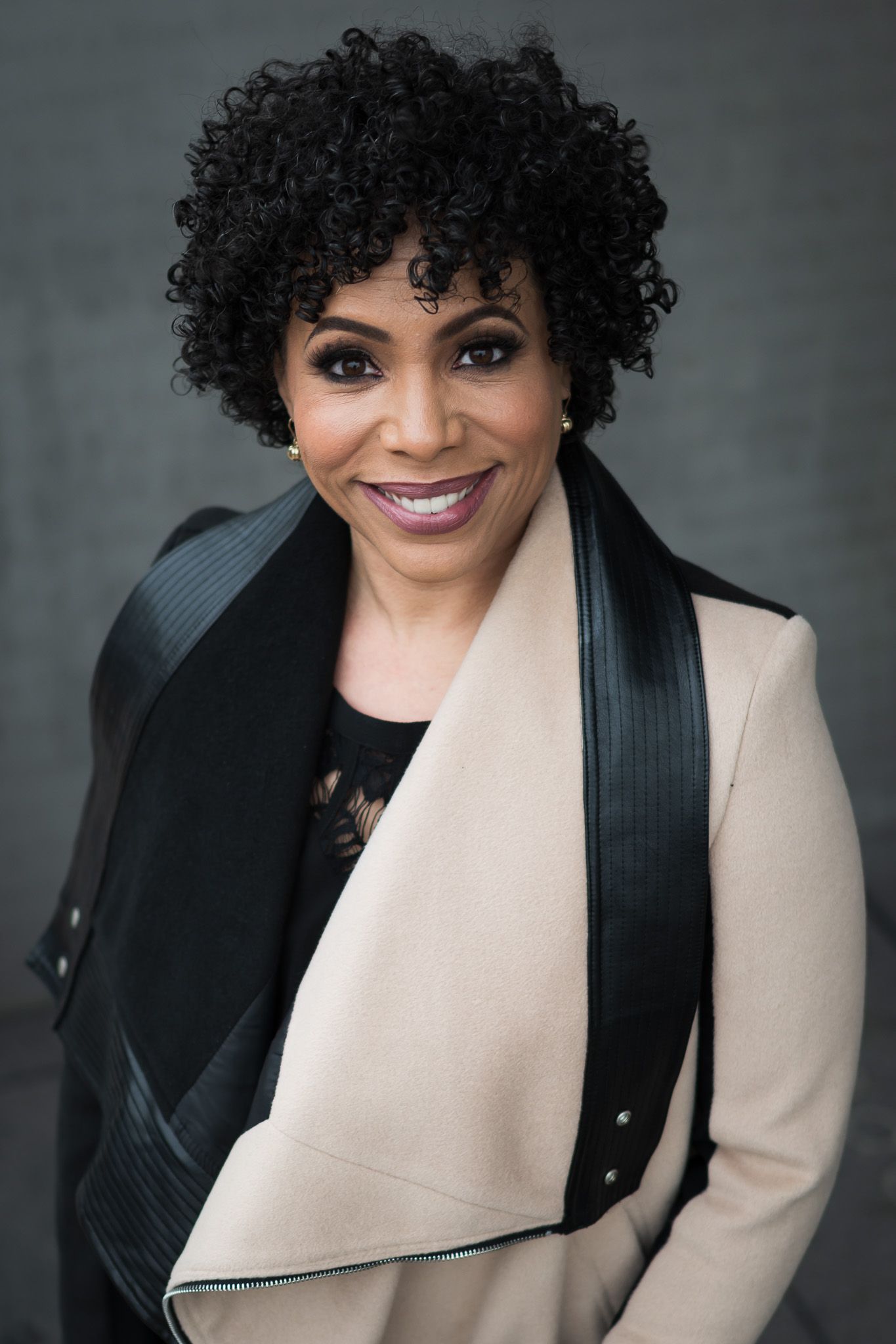 A smiling Black woman with curly hair wearing a beige blazer with black lapels poses for a professional portrait.