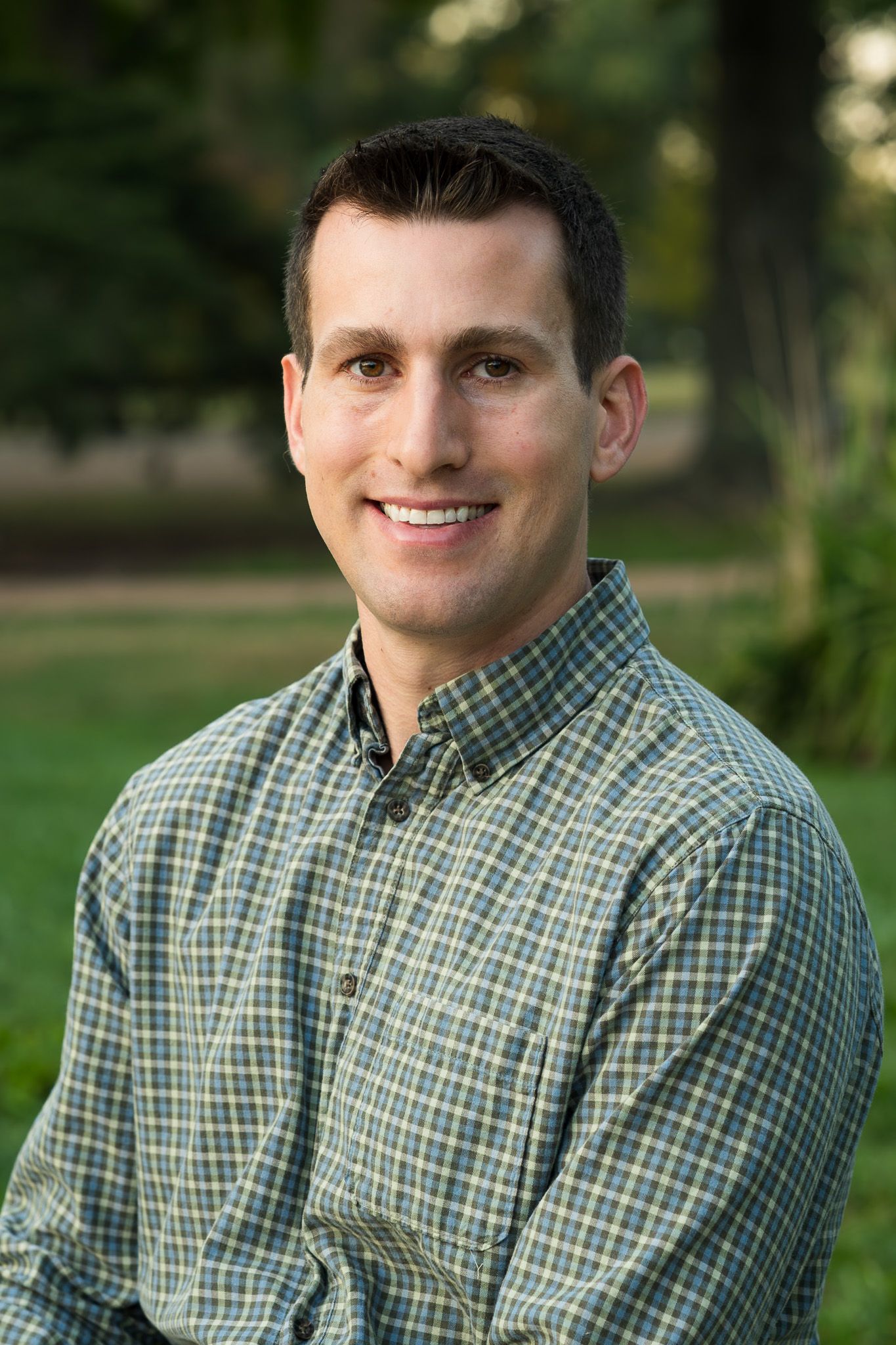 Smiling man in plaid shirt standing outdoors with green grass background.