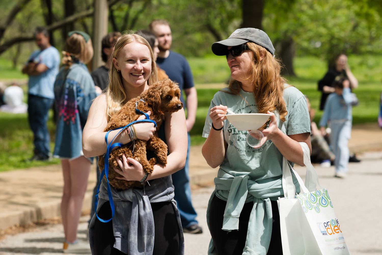 Two young women smile while walking in a park, one holding an adorable brown curly dog.