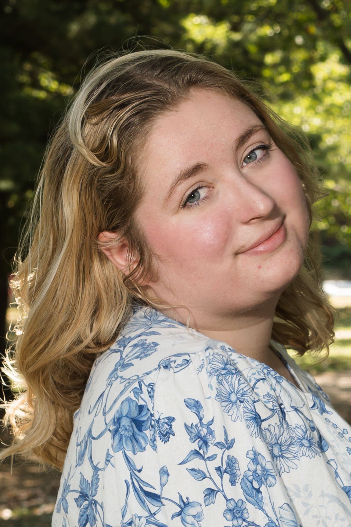 Young woman with blonde hair in blue floral dress smiling outdoors with green trees in background.