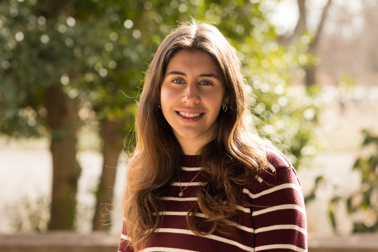 Young woman with long brown hair smiles wearing striped sweater outdoors.