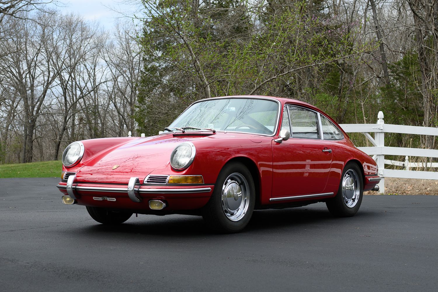Front three-quarter view of a classic red Porsche 911 coupe near a white fence with spring trees.