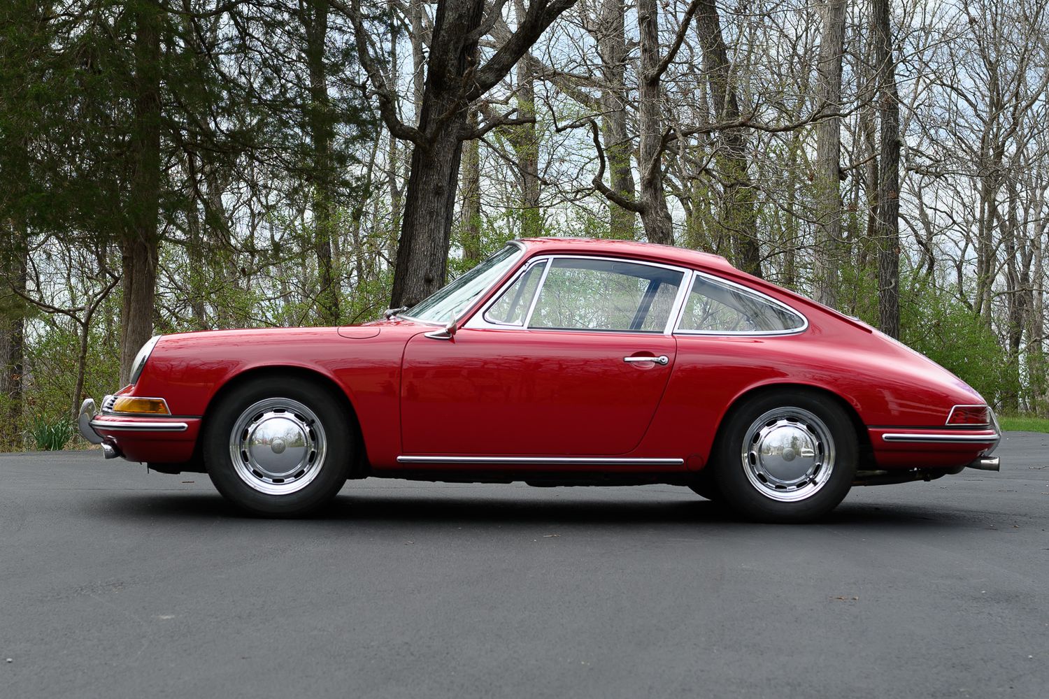 Side profile of a vintage red Porsche 911 coupe parked on a paved driveway with trees behind.