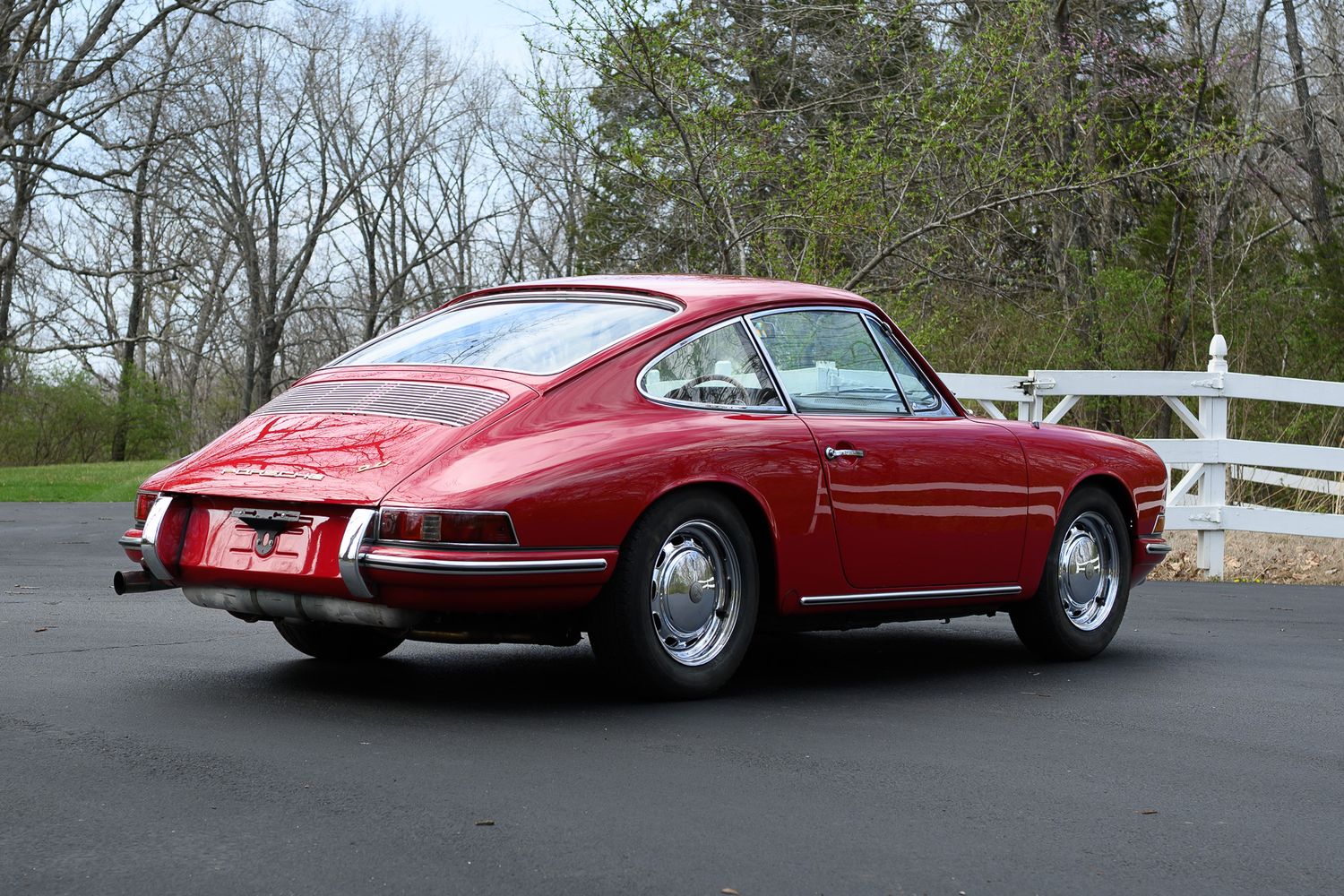Rear three-quarter view of a classic red Porsche 911 coupe near a white wooden fence outdoors.