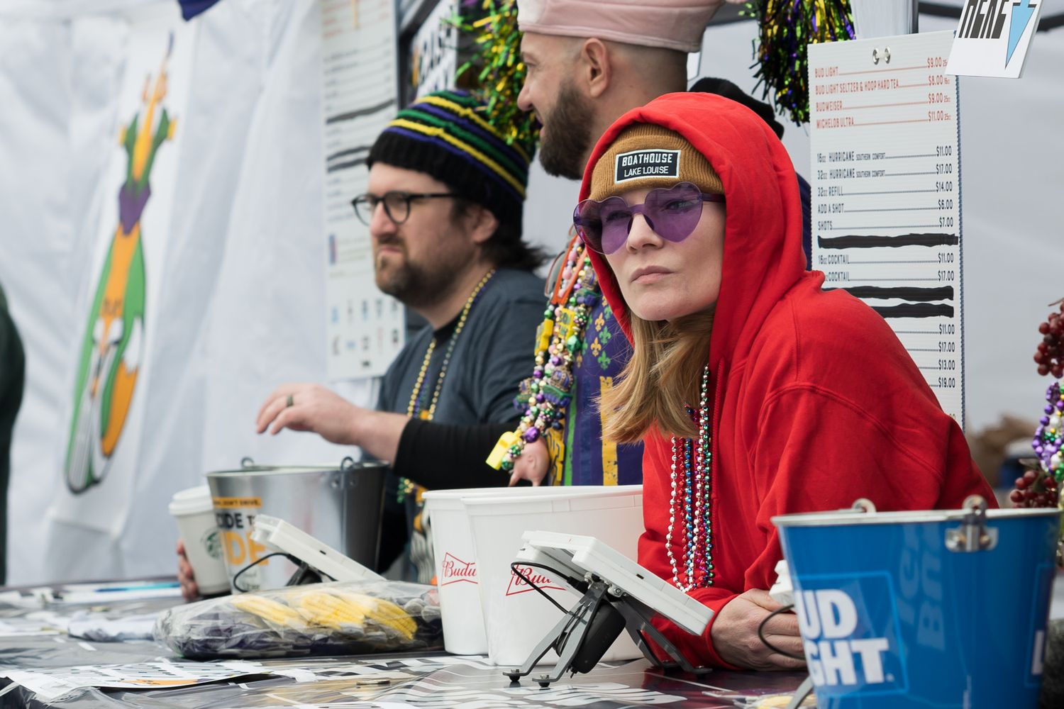 People working at an outdoor festival booth decorated with Mardi Gras beads and colorful signs.