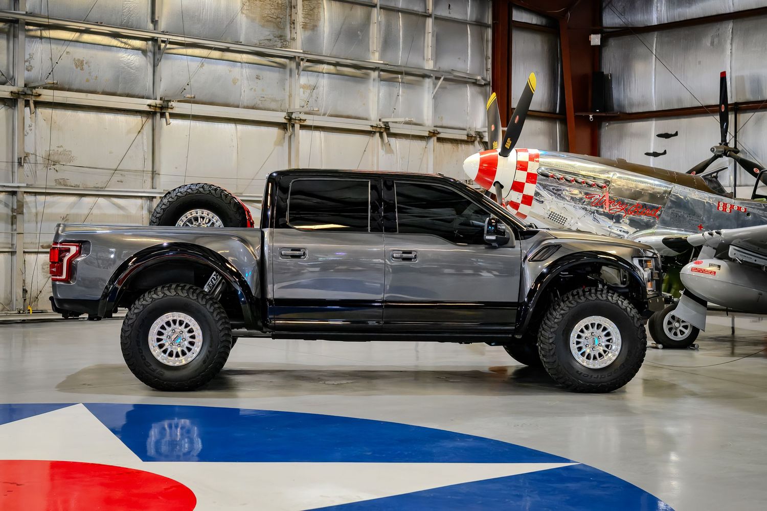 A lifted gray Ford F-150 Raptor on white wheels displayed in an aviation hangar beside a vintage WWII fighter plane.