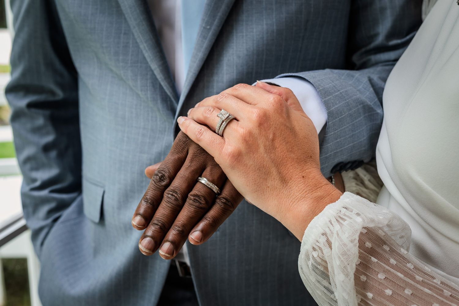 Interracial couple holding hands showing wedding rings, man in gray suit, woman in white lace dress, close-up.