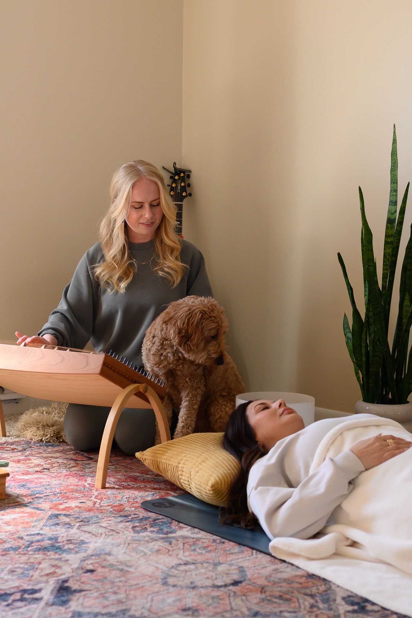 Blonde woman plays dulcimer while brown doodle dog sits nearby and woman relaxes on floor.