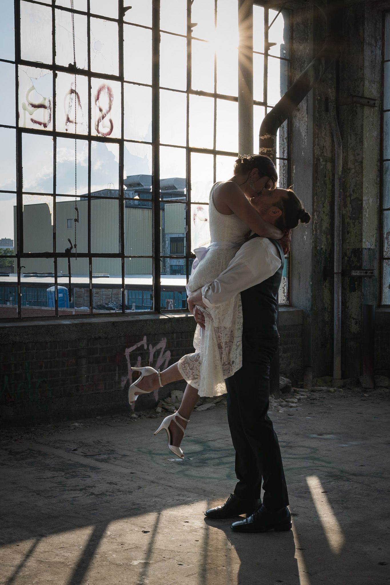 Groom lifts bride in a sunlit abandoned warehouse in a romantic wedding photo.