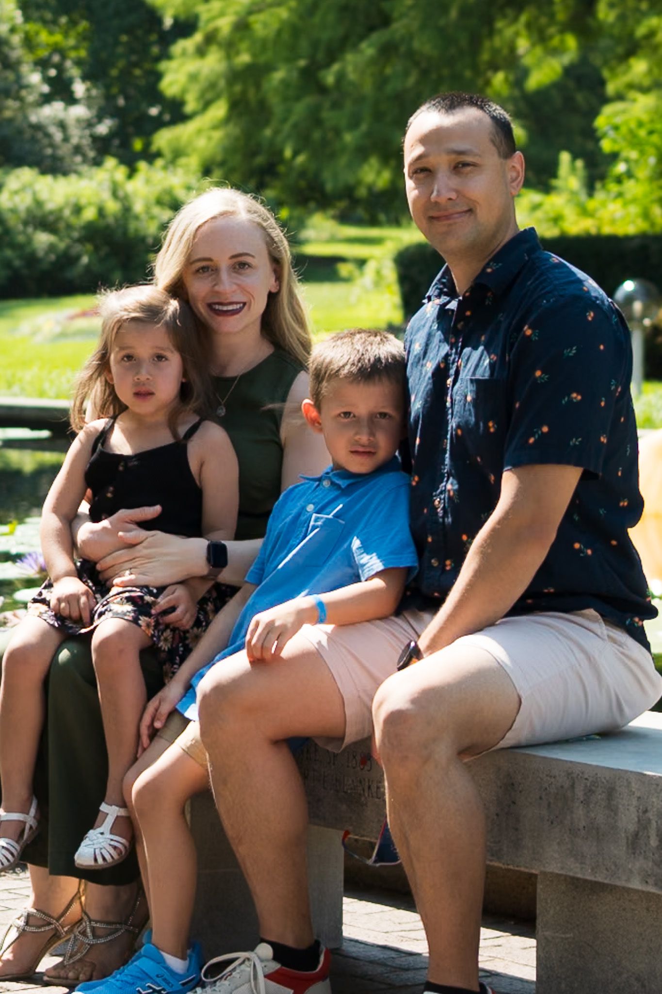 Happy family of four posing outdoors on a bench in a lush green park on a sunny summer day.