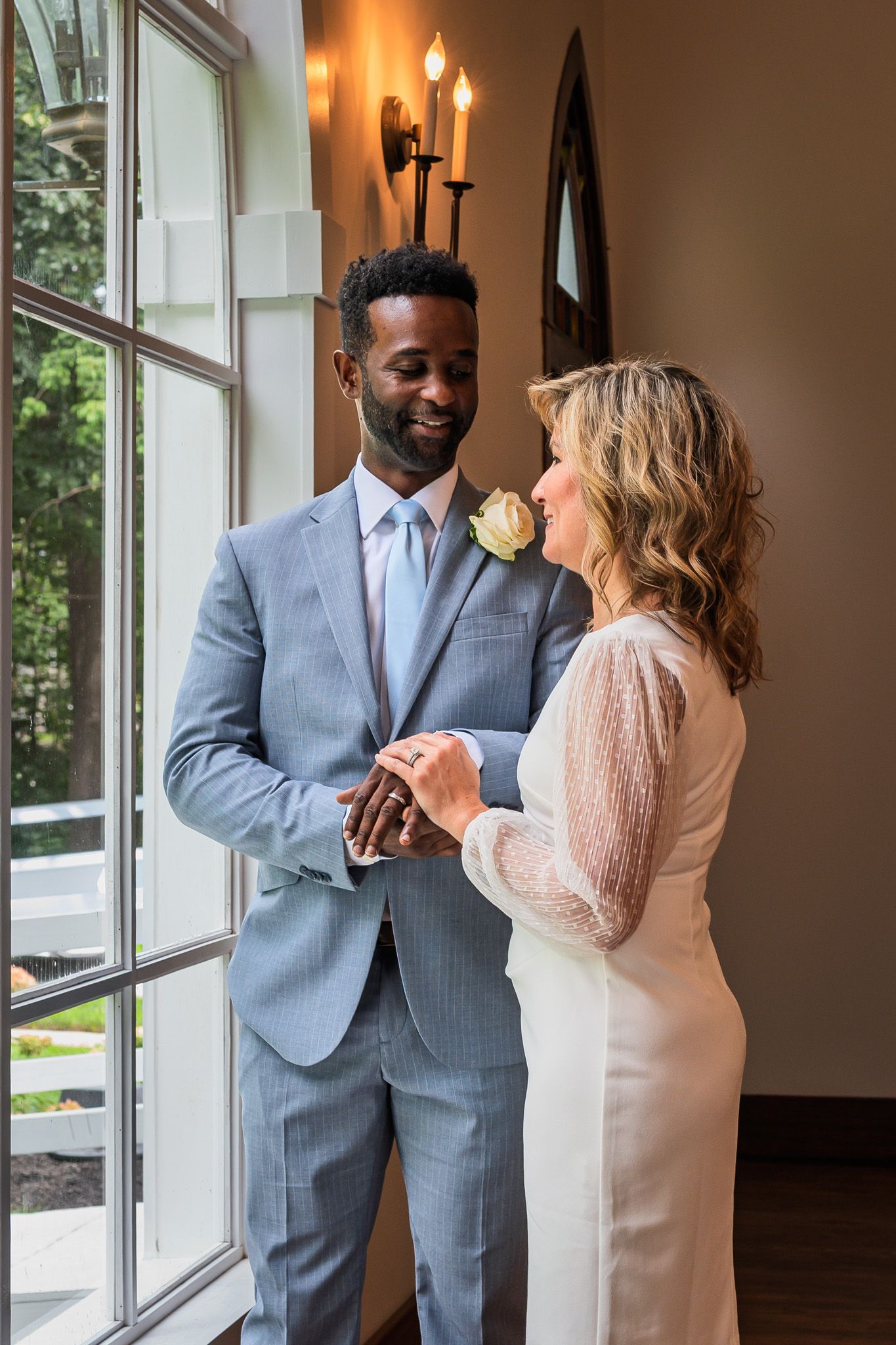 A bride and groom hold hands and smile at each other near a window, groom in a light blue suit with a white boutonniere.
