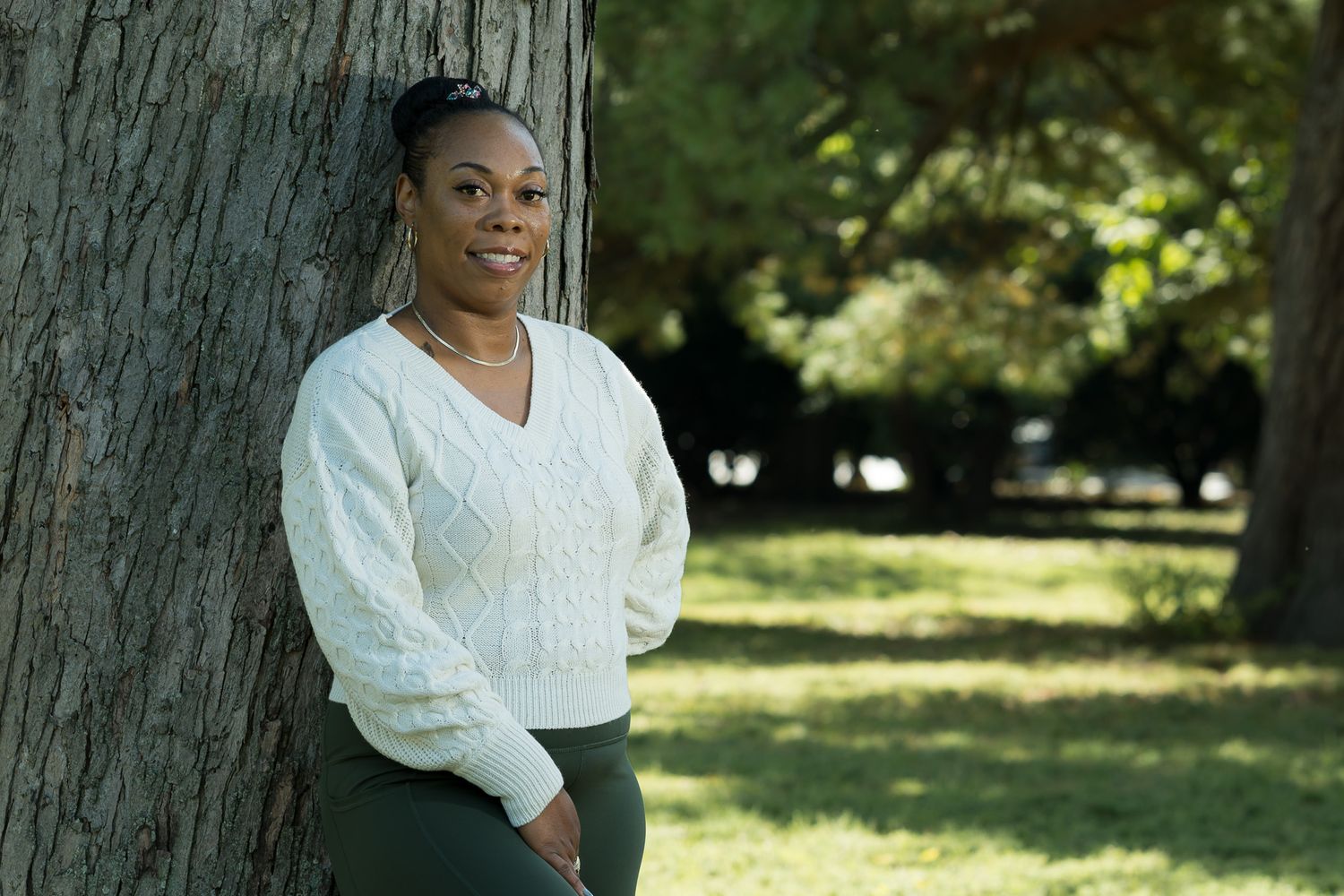 A Black woman in a white sweater leans against a tree trunk in a sunlit park, smiling calmly.