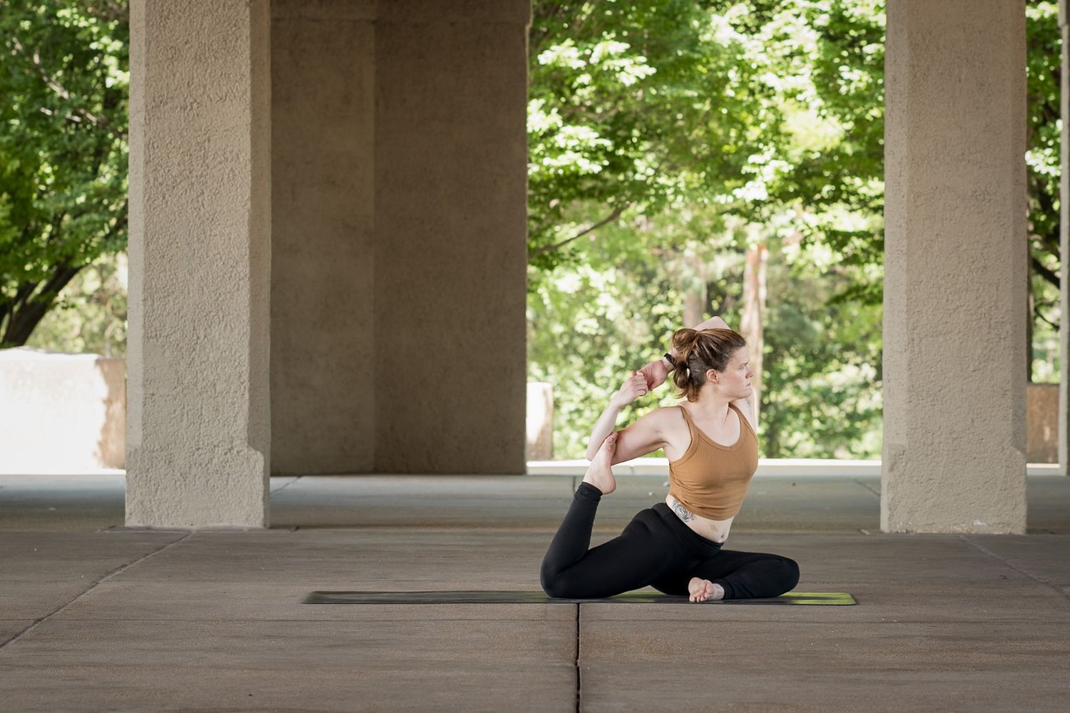 Woman practicing king pigeon yoga pose on mat under concrete pillars with green trees in background.