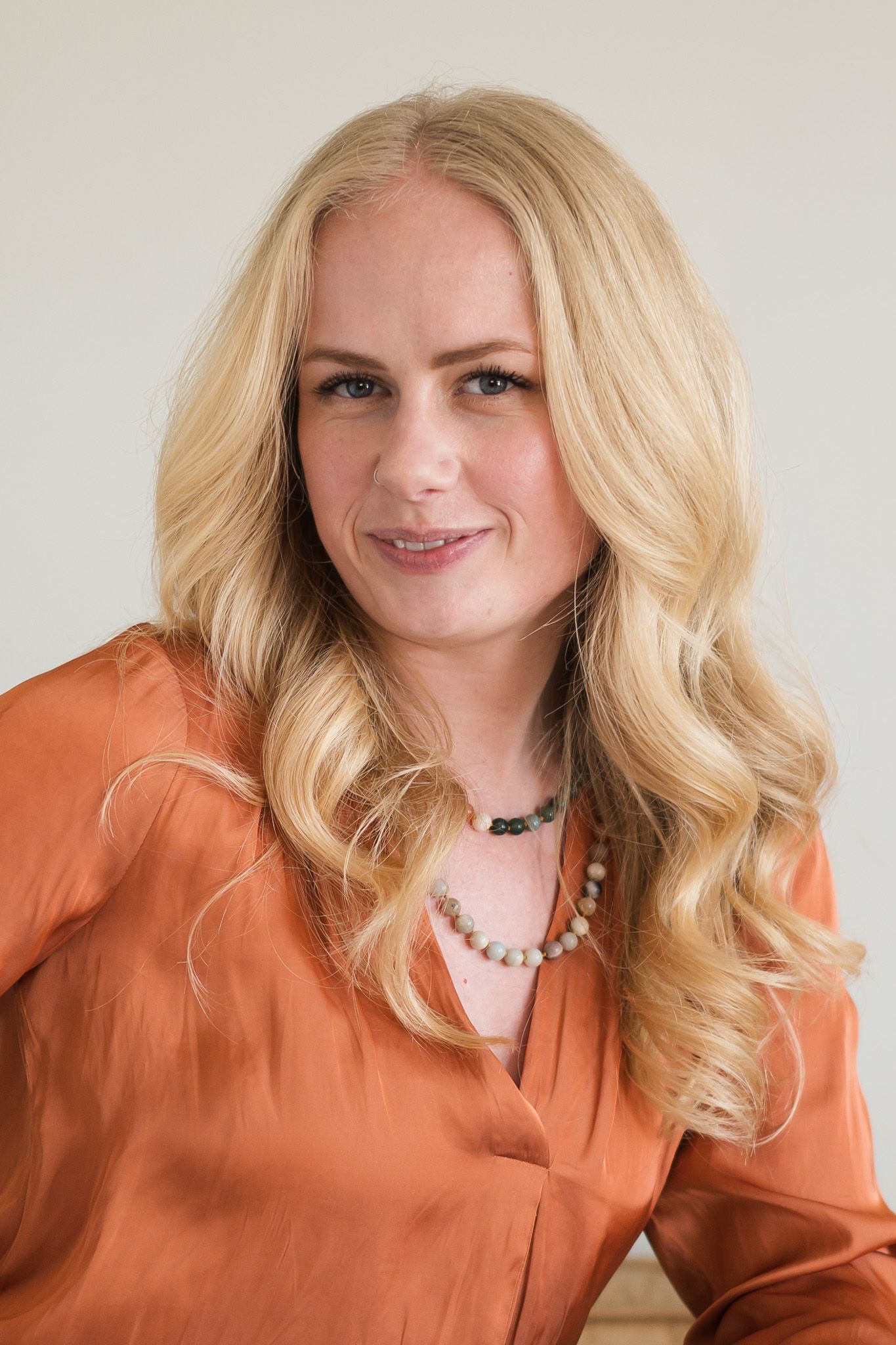 Professional portrait of smiling blonde woman in rust orange blouse with layered necklace.