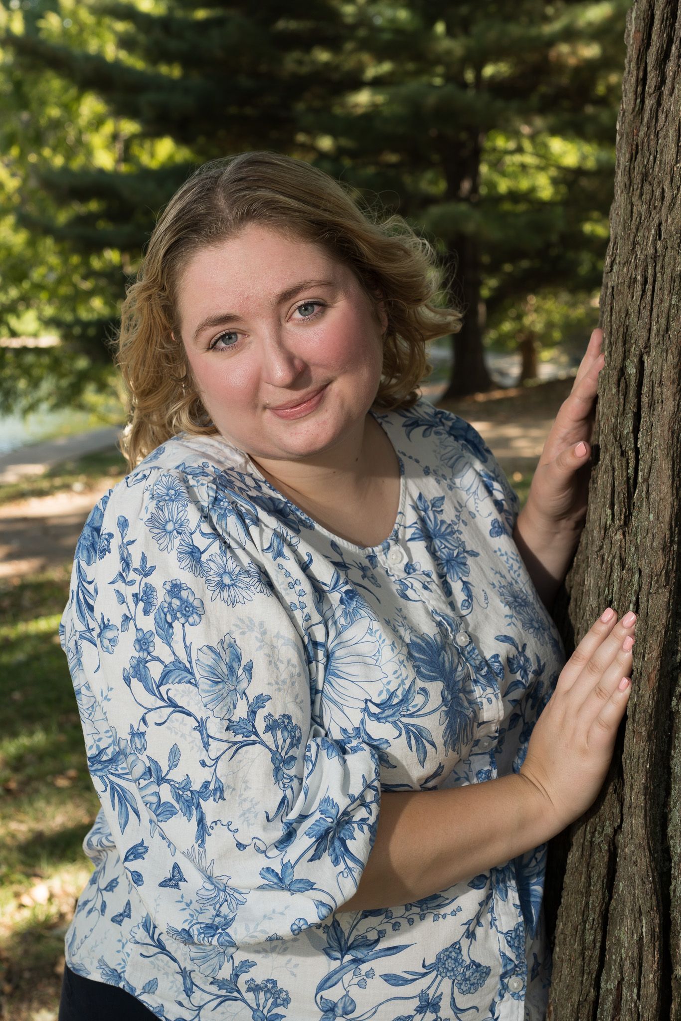 Young blonde woman in blue floral blouse leans against tree in sunny park.