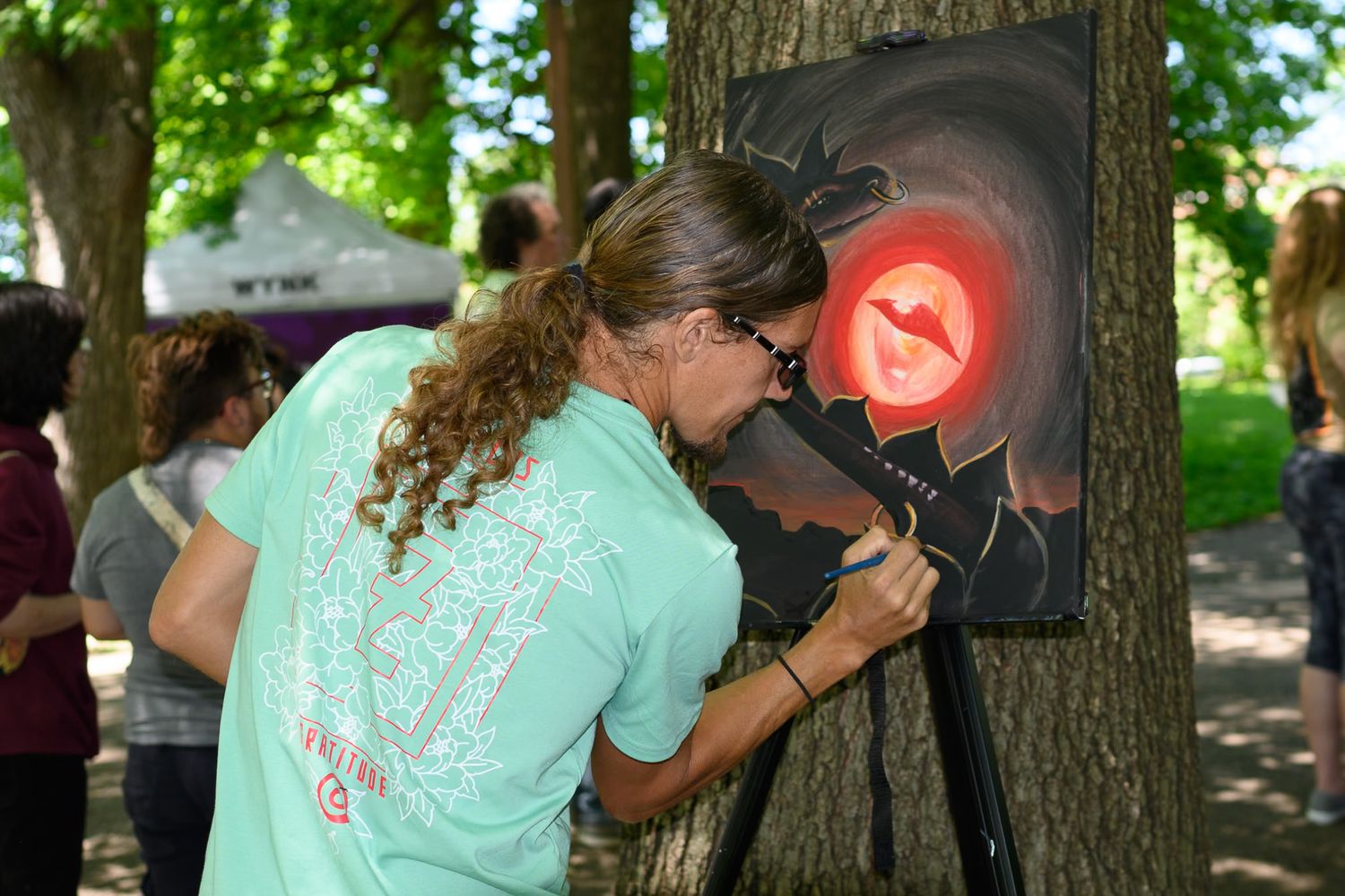 Artist with long hair painting a glowing red circular design on canvas at an outdoor art festival.