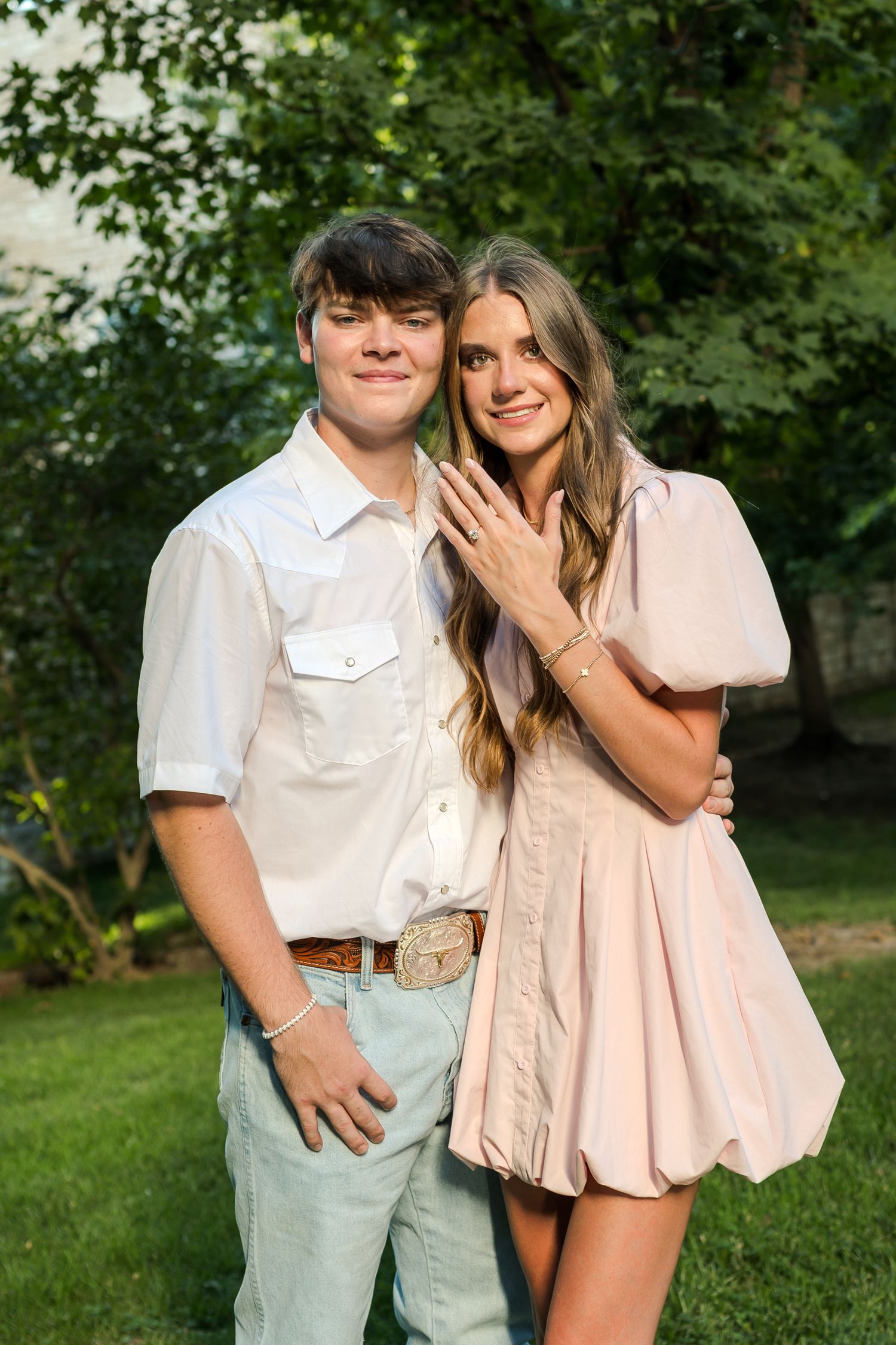 Happy young couple posing outdoors, woman showing engagement ring, wearing pastel outfits with green trees in background.