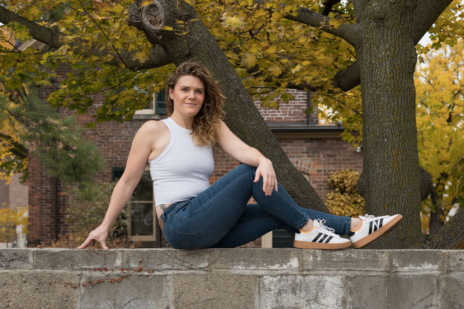 Young woman in white tank top and jeans posing on stone wall with fall foliage.
