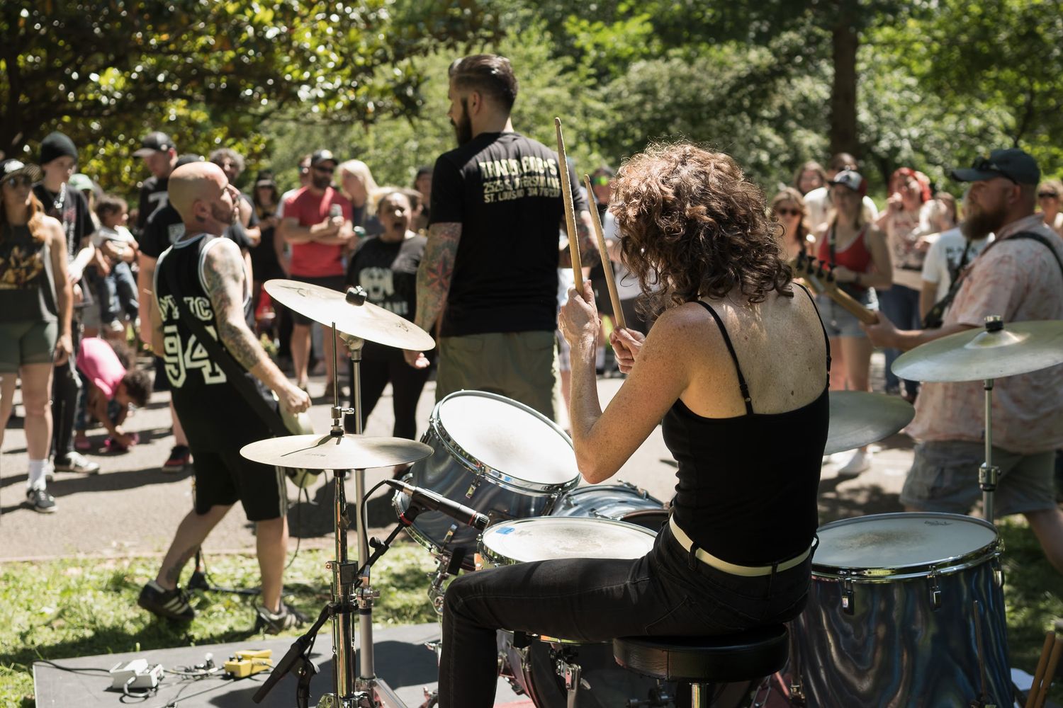 Female drummer performing outdoors at a park concert with crowd watching in sunny weather.