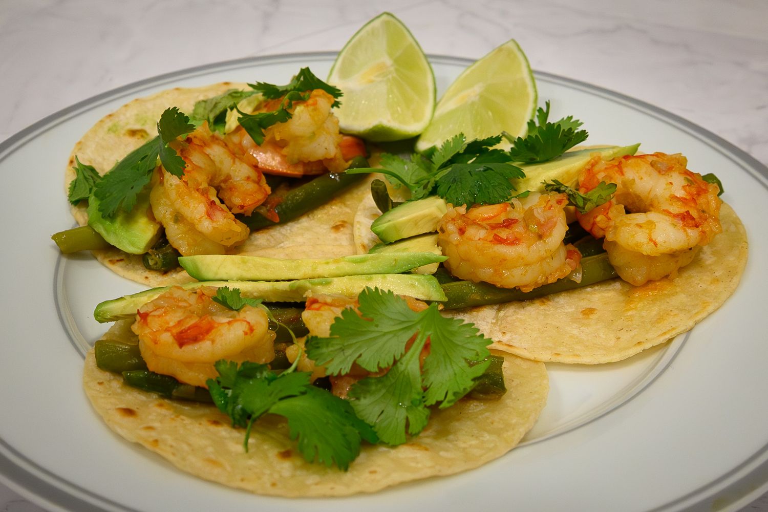 Shrimp tacos on corn tortillas topped with avocado, cilantro, and lime wedges on a white plate.