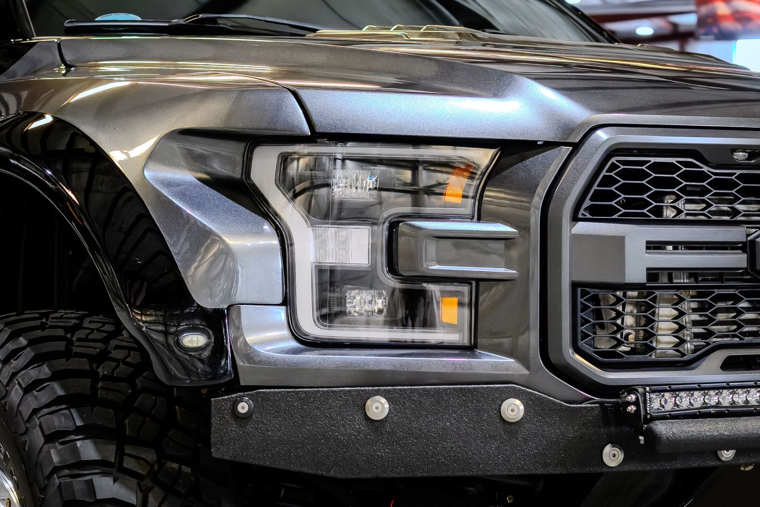 Close-up of a Ford F-150 Raptor front headlight and grille, showcasing aggressive styling on a dark gray truck.