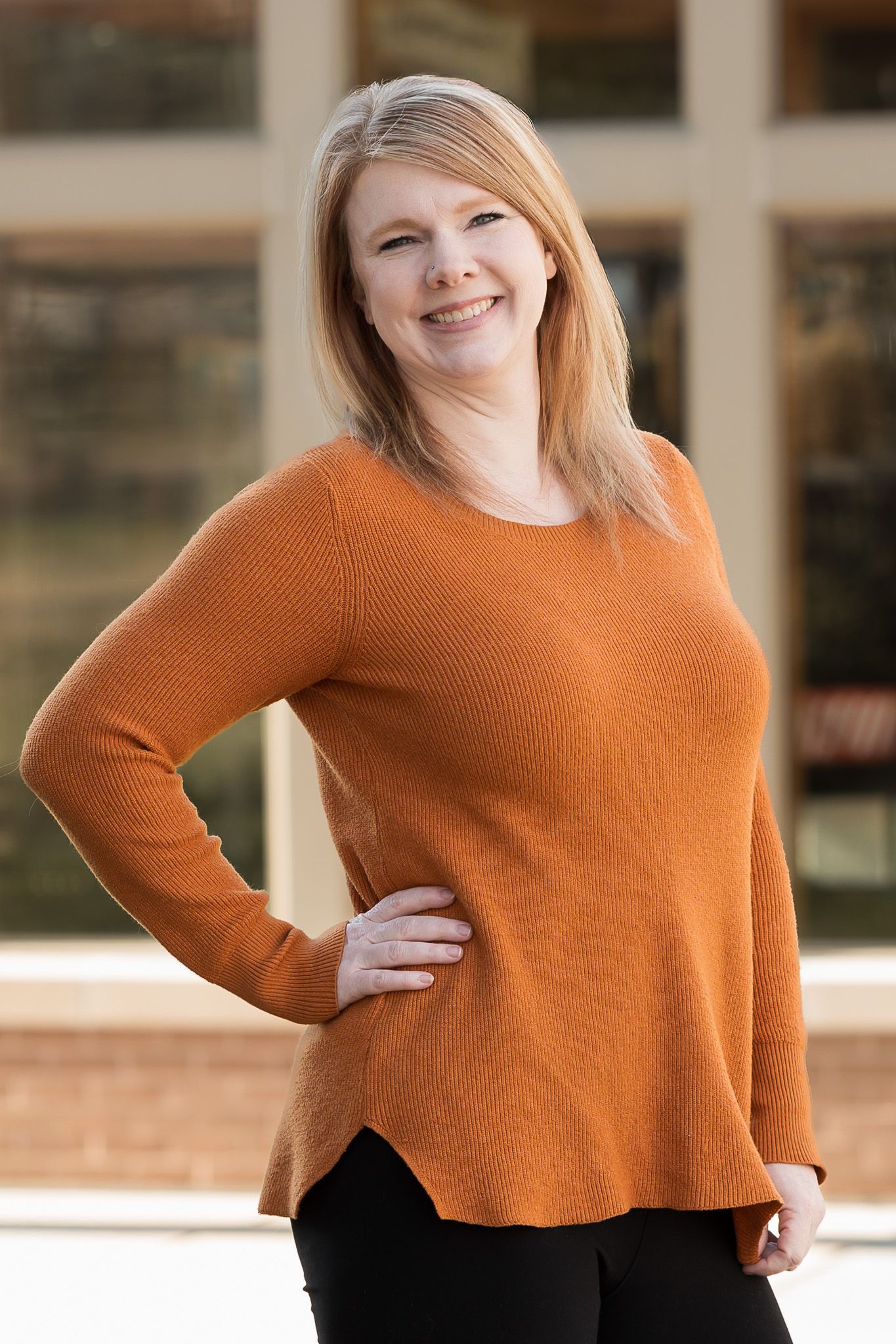 Smiling blonde woman in rust orange knit sweater posing with hand on hip outdoors.