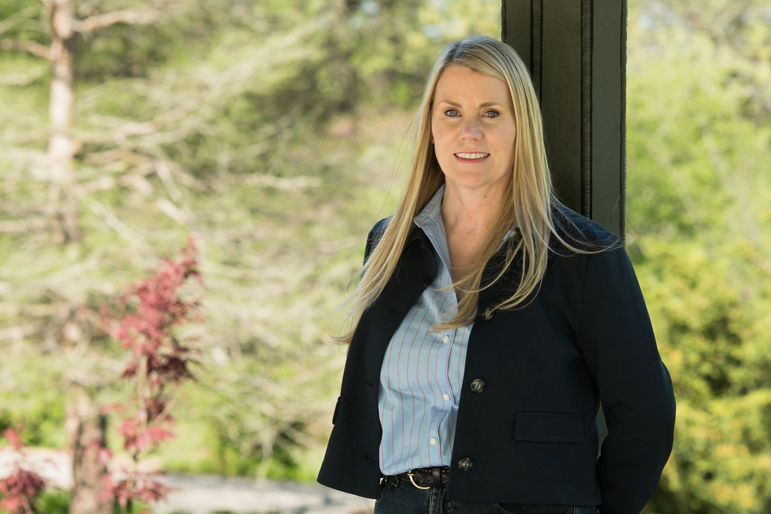 Professional woman in navy blazer smiling outdoors with lush green trees in background.