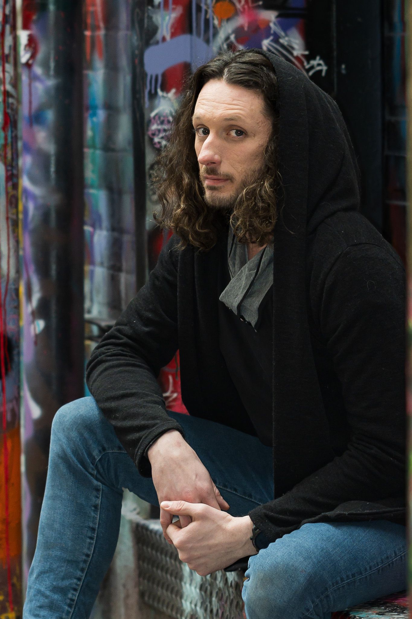 Man with long curly hair in black hoodie sitting against colorful graffiti wall in urban setting.