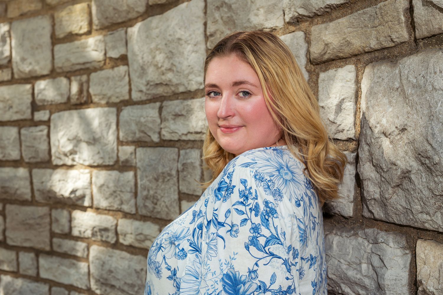 Young blonde woman in blue floral blouse leaning against rustic stone wall smiling.