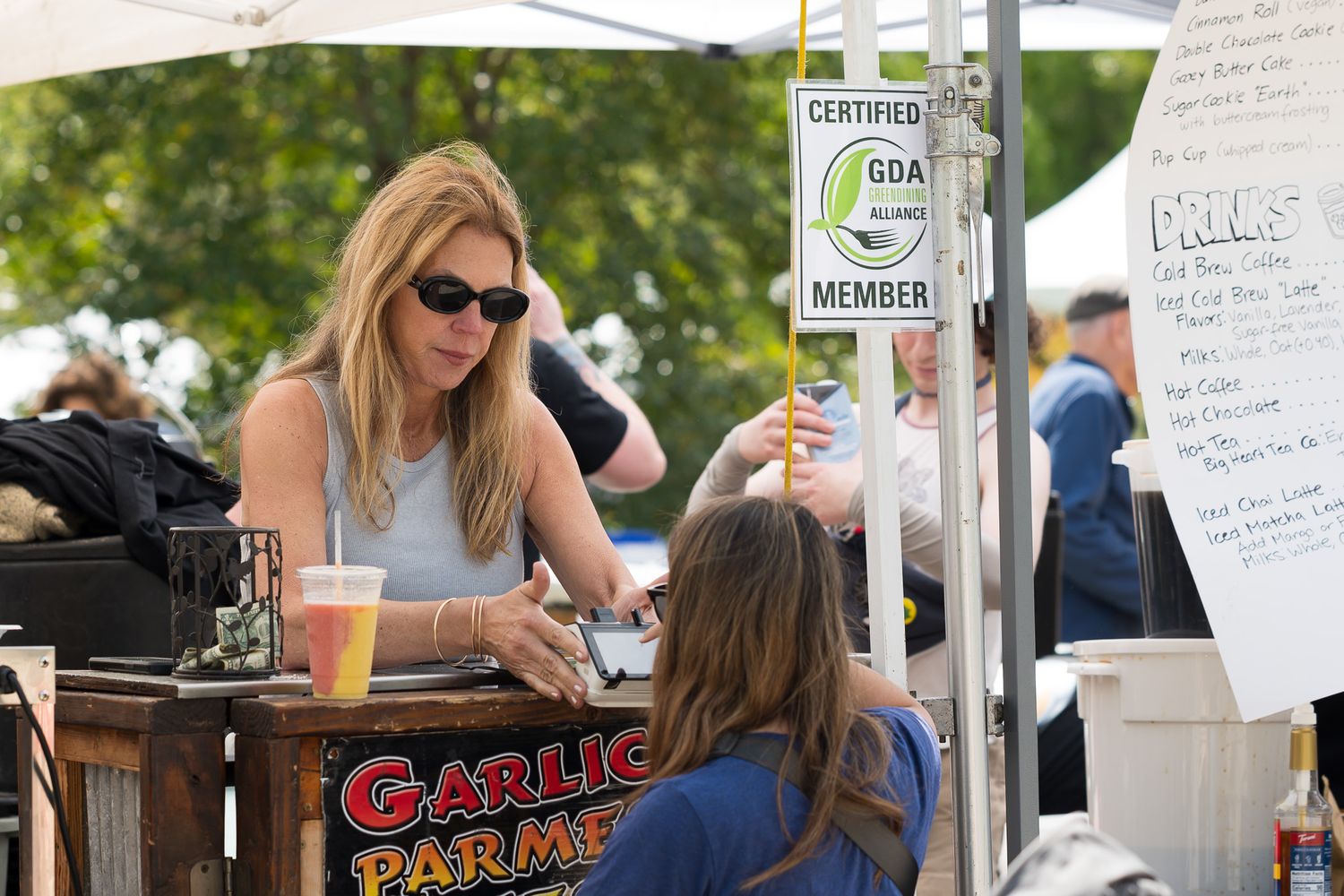 Blonde woman in sunglasses at outdoor food festival booth with Garlic Parmesan sign and GDA member certification.