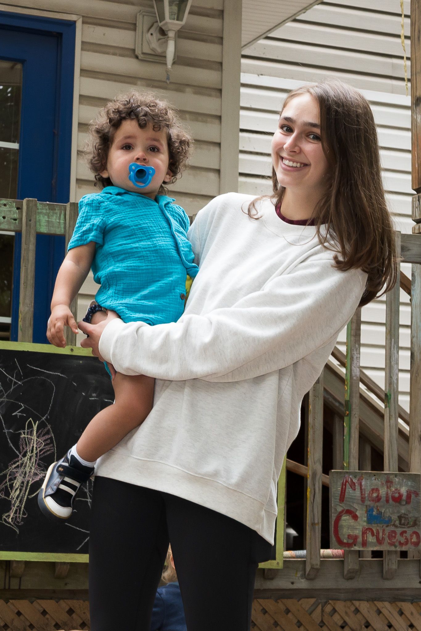 Young woman smiling while holding a toddler with a blue pacifier outside a house.
