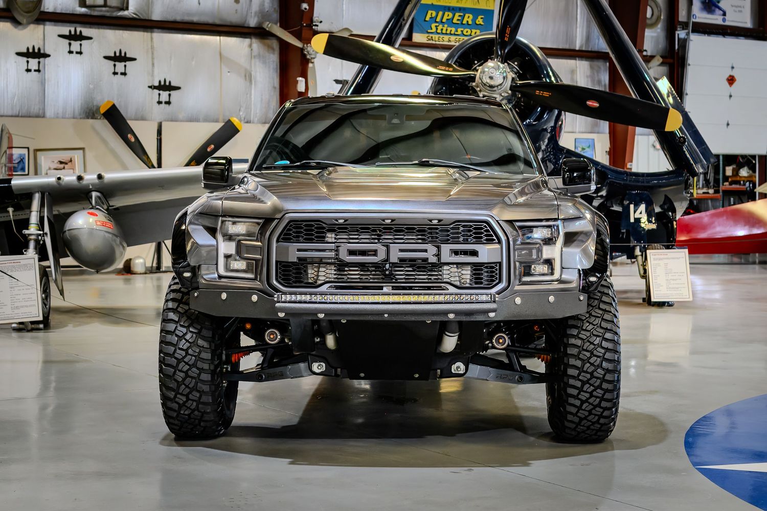 Front view of a lifted Ford Raptor truck with off-road tires displayed inside an aviation hangar.