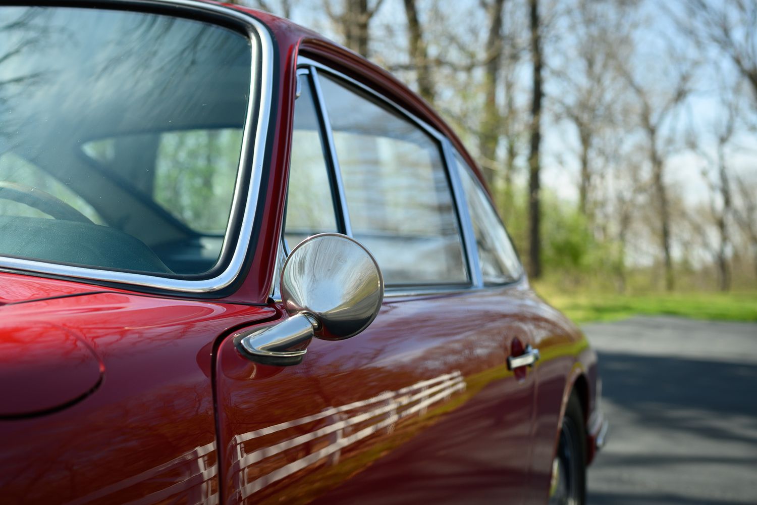 Close-up of a vintage red Porsche 911 door mirror and window trim with trees in background.