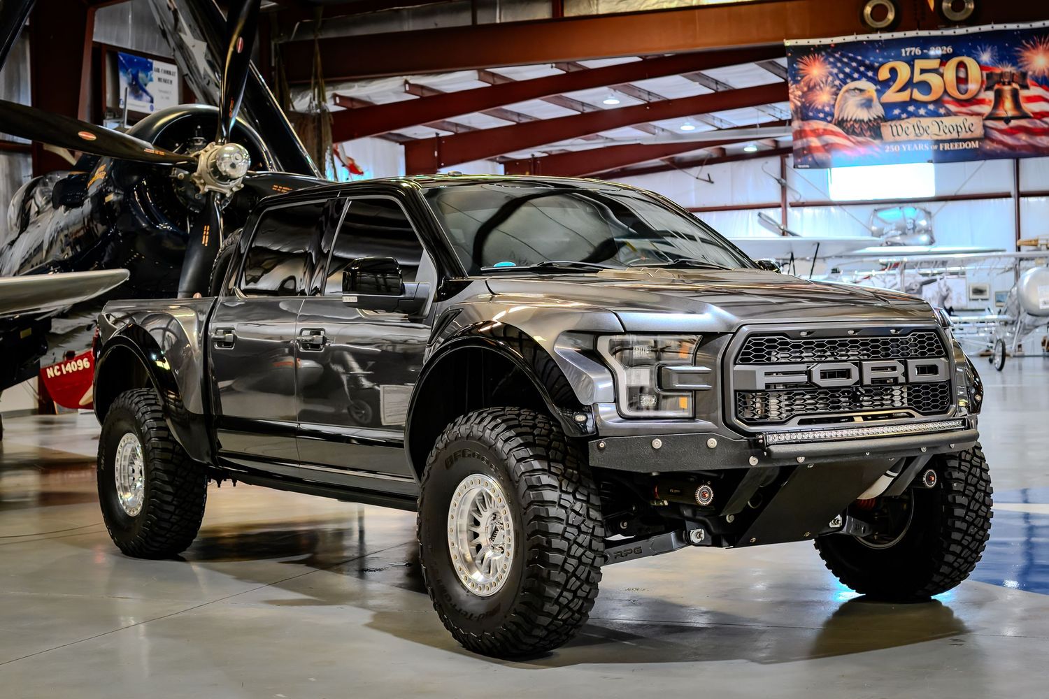 Modified Ford Raptor off-road truck with aggressive stance and large tires displayed inside an aviation hangar.