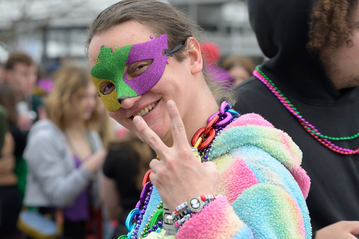 Young woman wearing colorful rainbow hoodie and glittery Mardi Gras mask making peace sign at festive outdoor celebration.